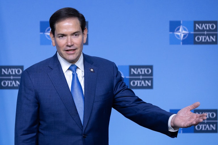 Marco Rubio smiles while speaking and stretches out his hand whlie standing in front of a NATO backdrop.