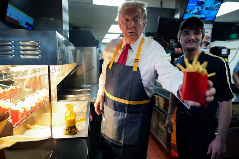 Donald Trump, wearing a McDonald's apron and standing by a fryer, reaches out holding a container of french fries.
