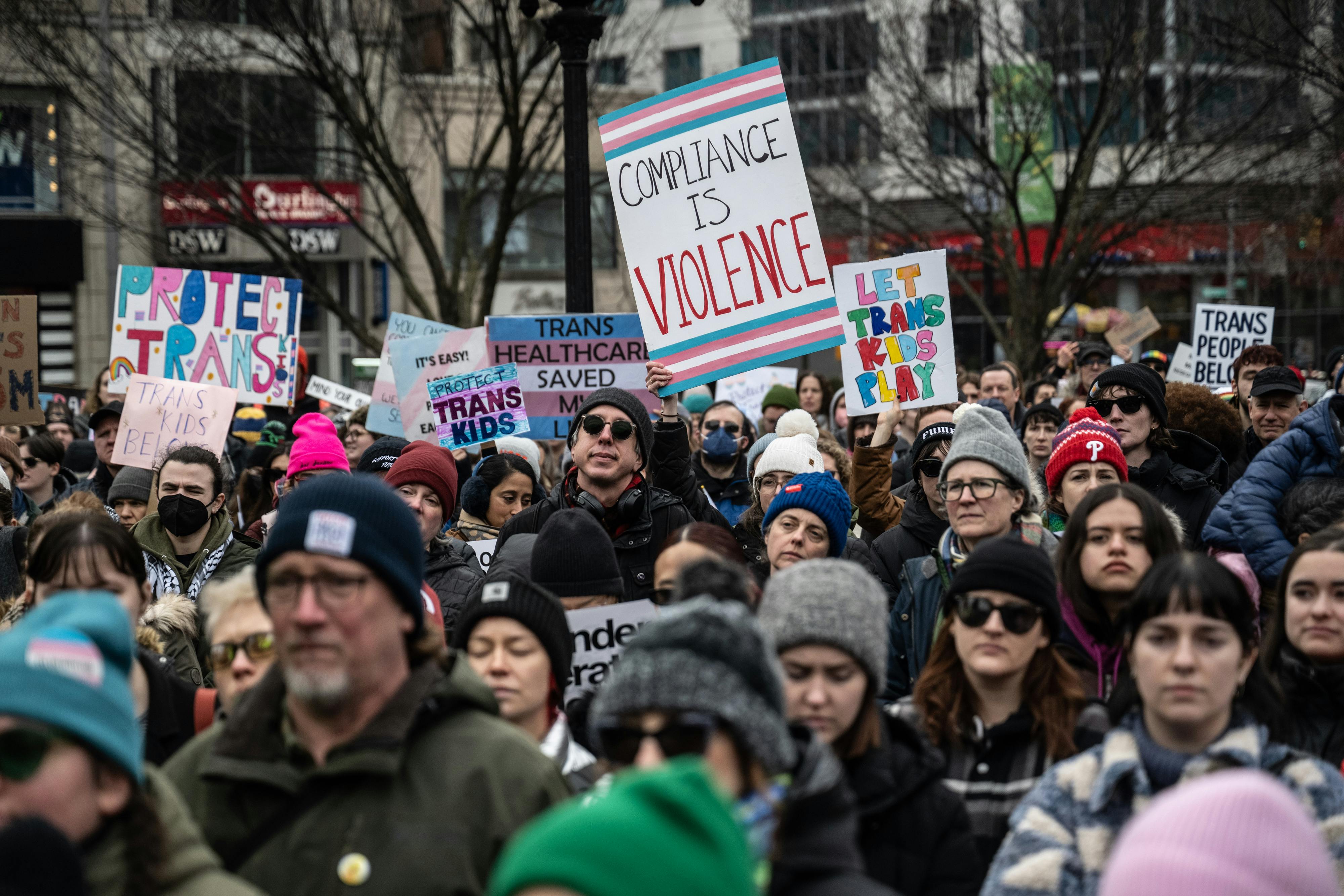 Demonstrators hold signs reading "Compliance Is Violence" and "Protect Trans Kids."