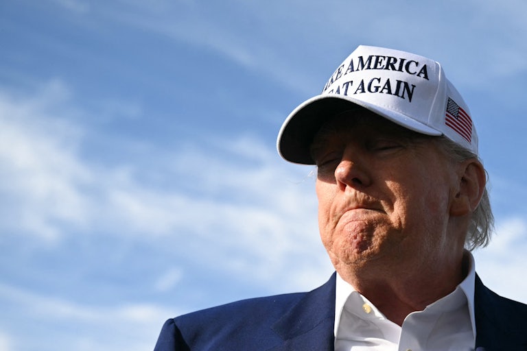Donald Trump purses his lips and looks down while wearing a white "Make America Great Again" hat