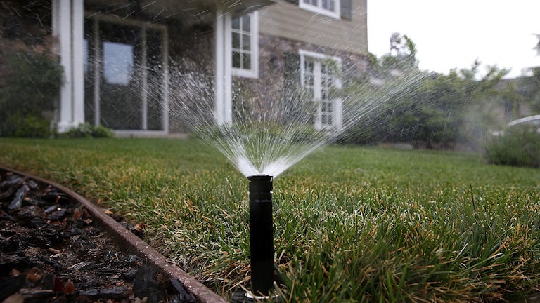 A sprinkler sprays water over a lawn with a house in the background.