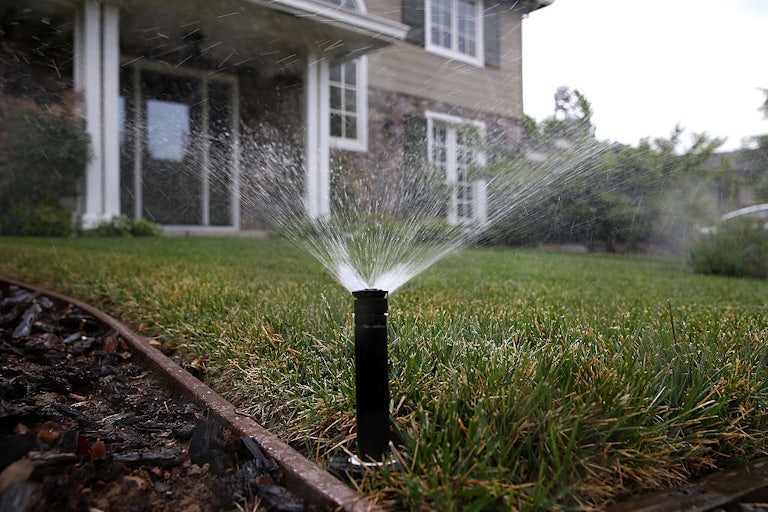 A sprinkler sprays water over a lawn with a house in the background.