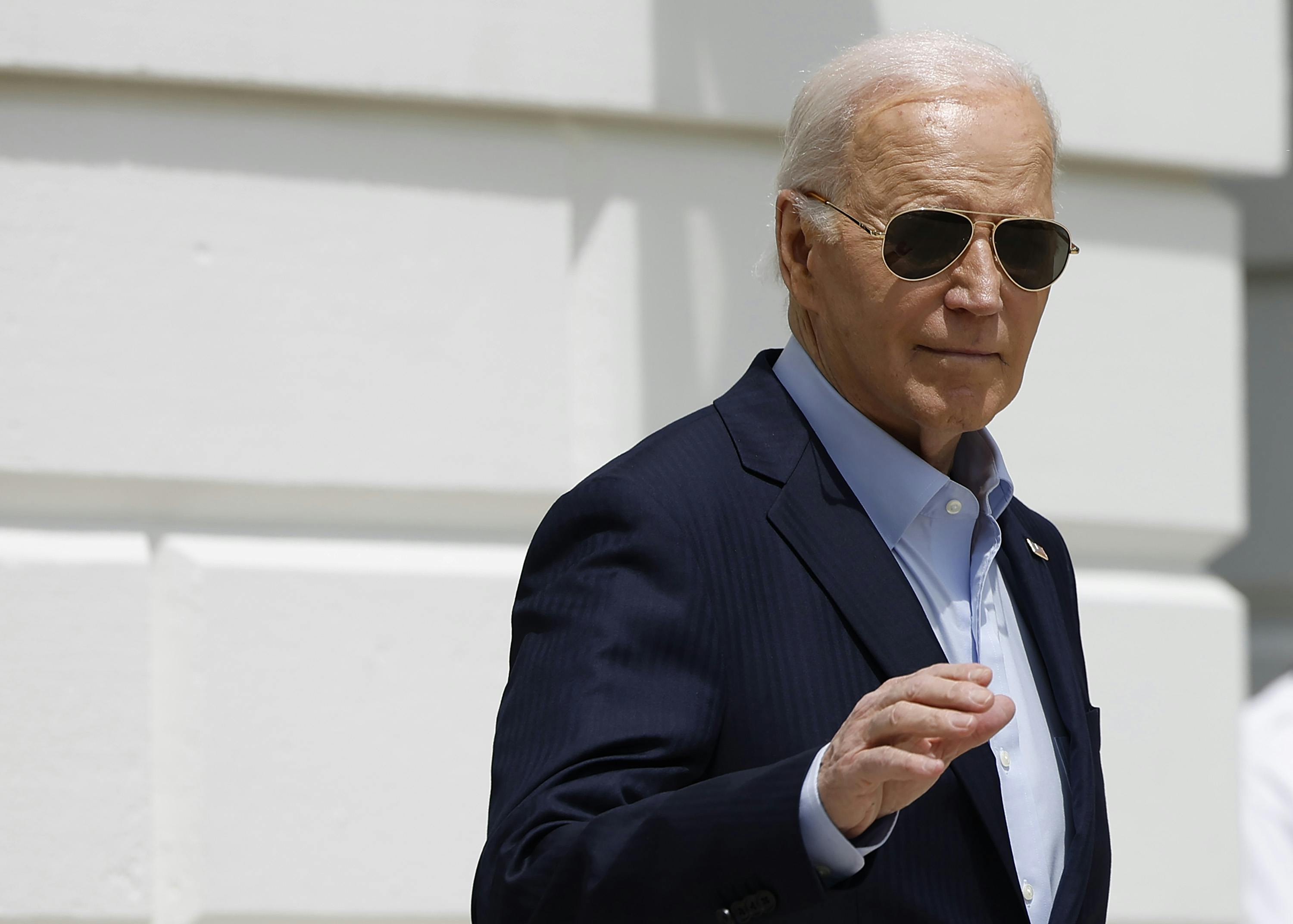 Joe Biden wears aviator sunglasses and raises his right hand as he leaves the White House on a sunny day. 