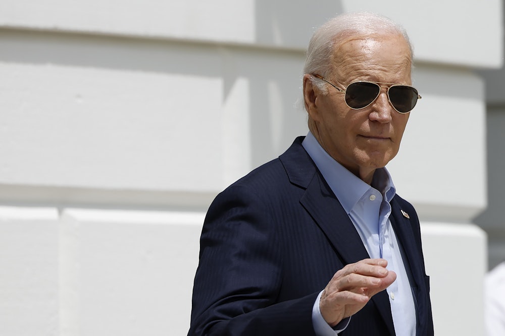 Joe Biden wears aviator sunglasses and raises his right hand as he leaves the White House on a sunny day.