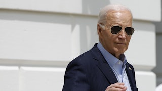 Joe Biden wears aviator sunglasses and raises his right hand as he leaves the White House on a sunny day.