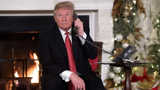 Donald Trump on the phone as he sits in front of the fireplace in a Christmas decorated room.