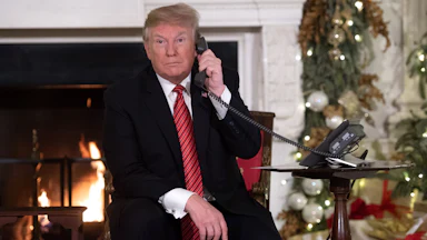 Donald Trump on the phone as he sits in front of the fireplace in a Christmas decorated room.