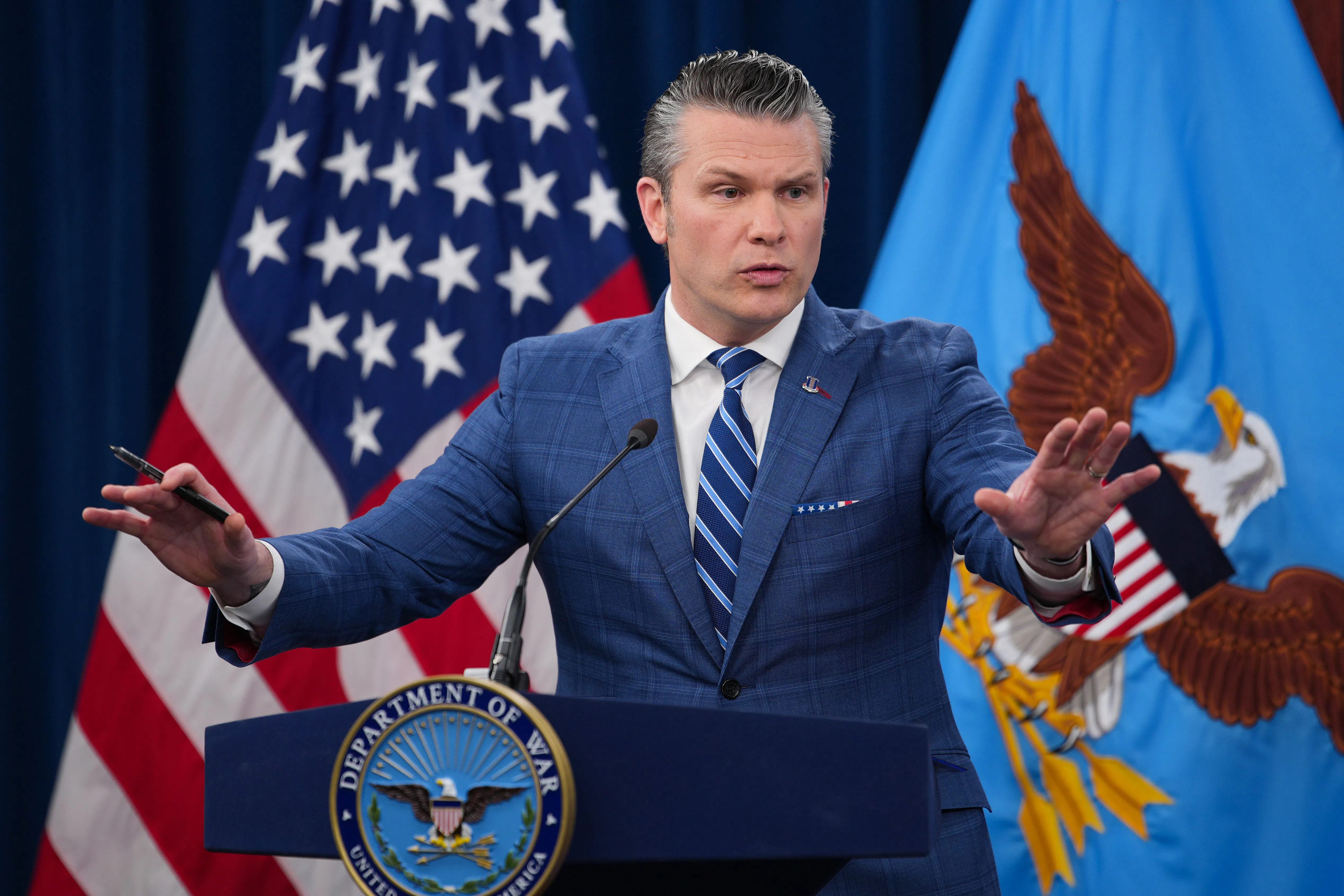 Defense Secretary Pete Hegseth holds both hands out while speaking at a podium