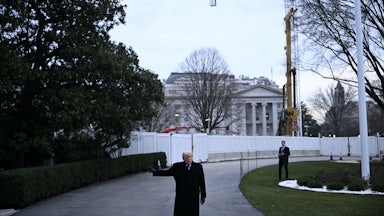 Donald Trump stands in front of the White House as a crane stands overhead.