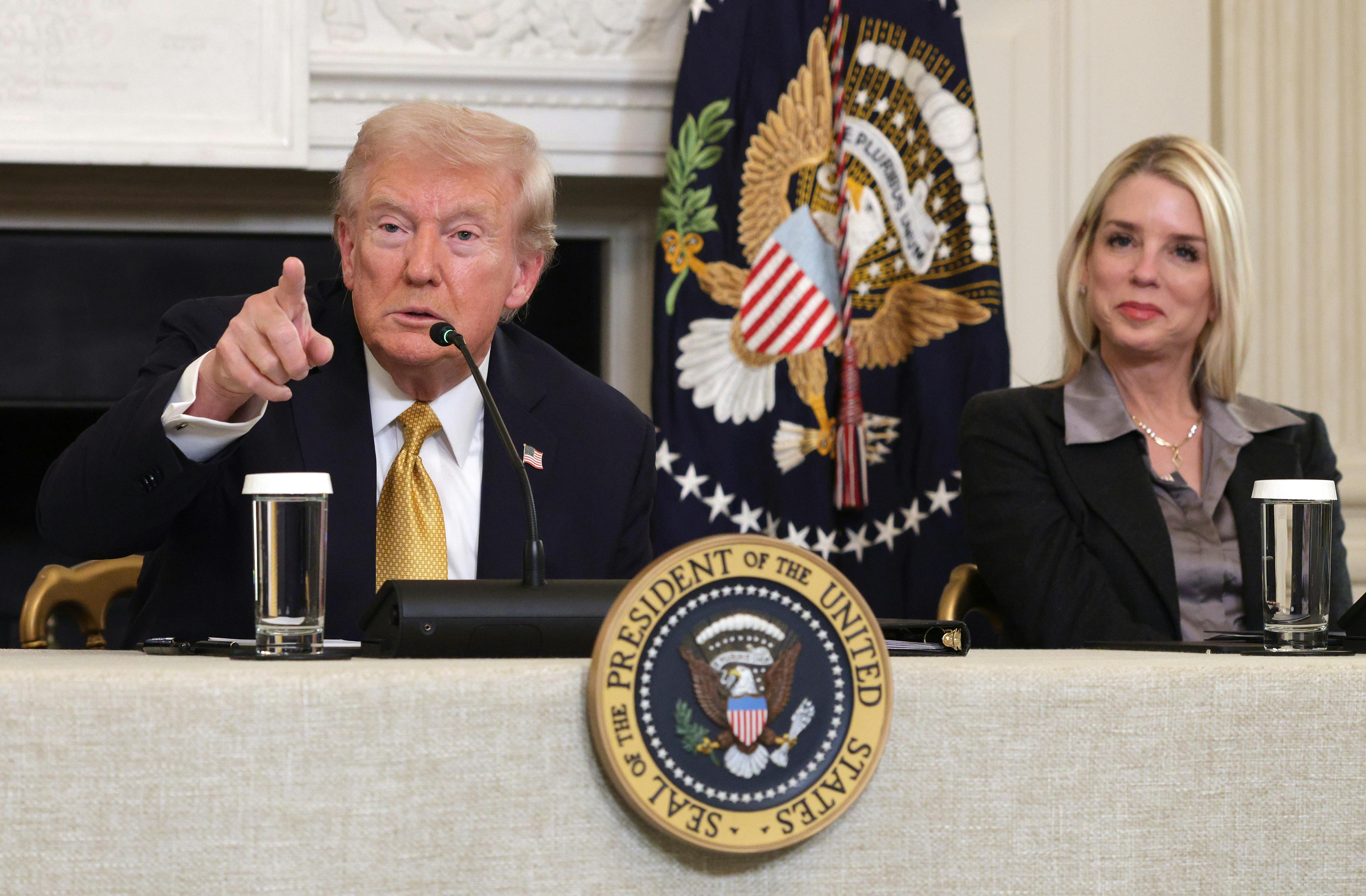 Donald Trump points as Attorney General Pam Bondi smiles. Both are seated a table with the presidential seal.