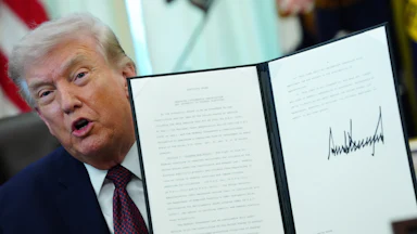 Donald Trump holds up a signed executive order and speaks while sitting at his desk in the Oval Office