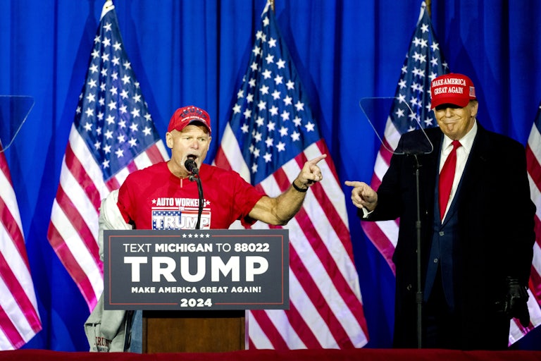 Brian Pannebecker speaks at a lectern while Donald Trump, wearing a MAGA hat, looks on and smiles