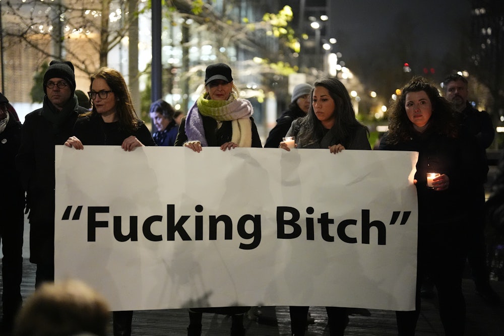 Standing outside in the dark, multiple people hold up a large poster reading "Fucking Bitch."