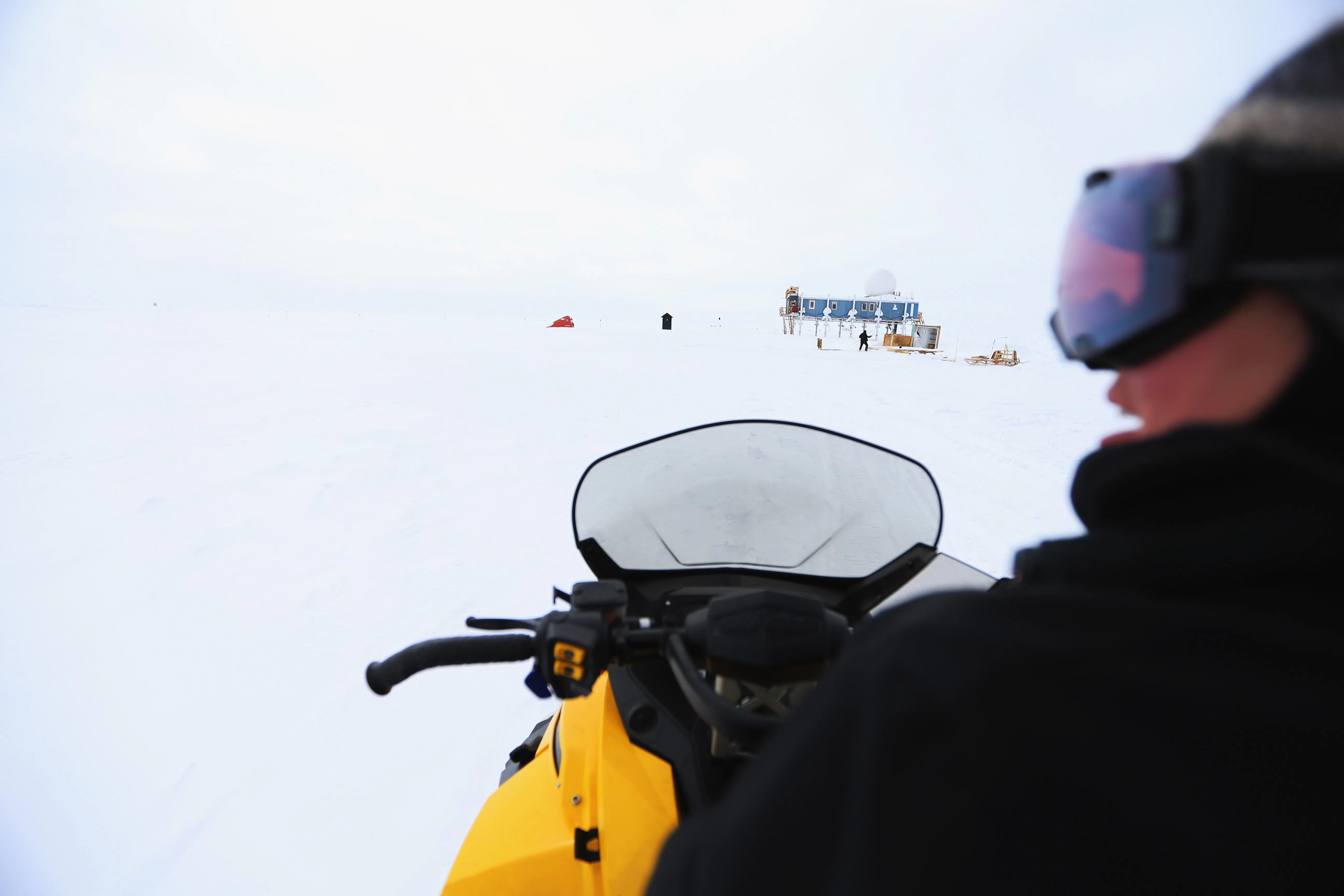 A person is seen on a snow vehicle with a small blue building on stilts in the background.
