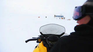 A person is seen on a snow vehicle with a small blue building on stilts in the background.