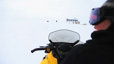 A person is seen on a snow vehicle with a small blue building on stilts in the background.