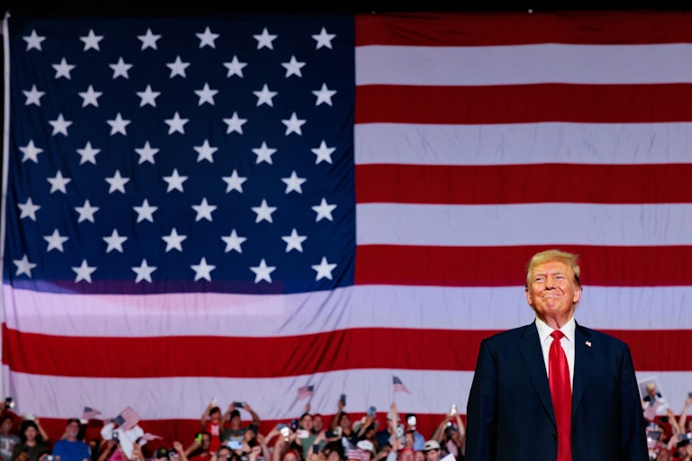 Donald Trump smiles as he stands in front of a giant American flag