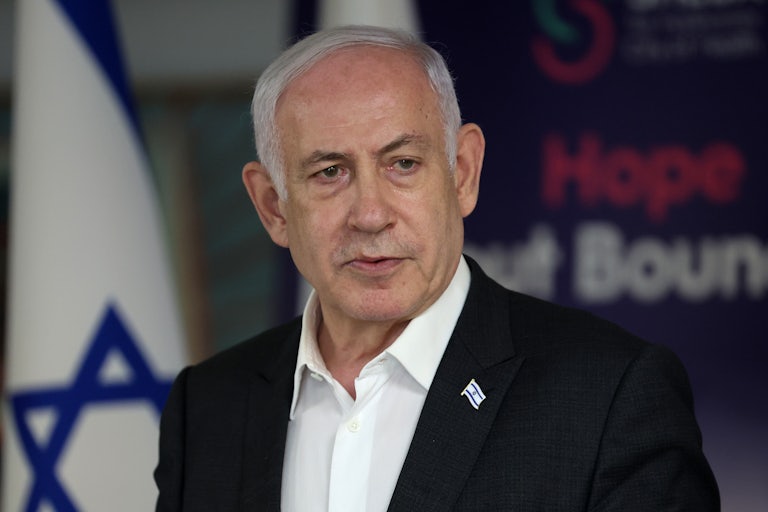 Photo of Israeli Prime Minister Benjamin Netanyahu looking slightly off-camera with a somewhat furrowed look on his face in front of an indeterminate sign and an Israeli flag.