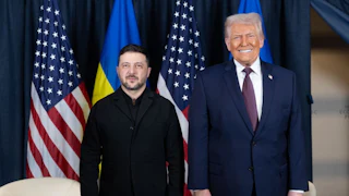 Volodymyr Zelenskiy and Donald Trump stand next to each other in front of Ukrainian and U.S. flags, with Trump smiling and Zelenskiy having a neutral expression.