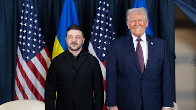 Volodymyr Zelenskiy and Donald Trump stand next to each other in front of Ukrainian and U.S. flags, with Trump smiling and Zelenskiy having a neutral expression.