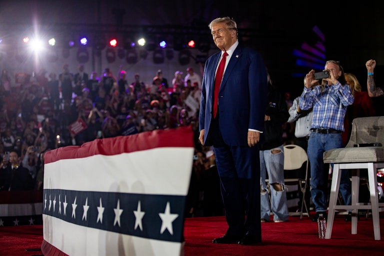 Donald Trump smiles at a campaign rally. Members of the crowd cheer and take photos.