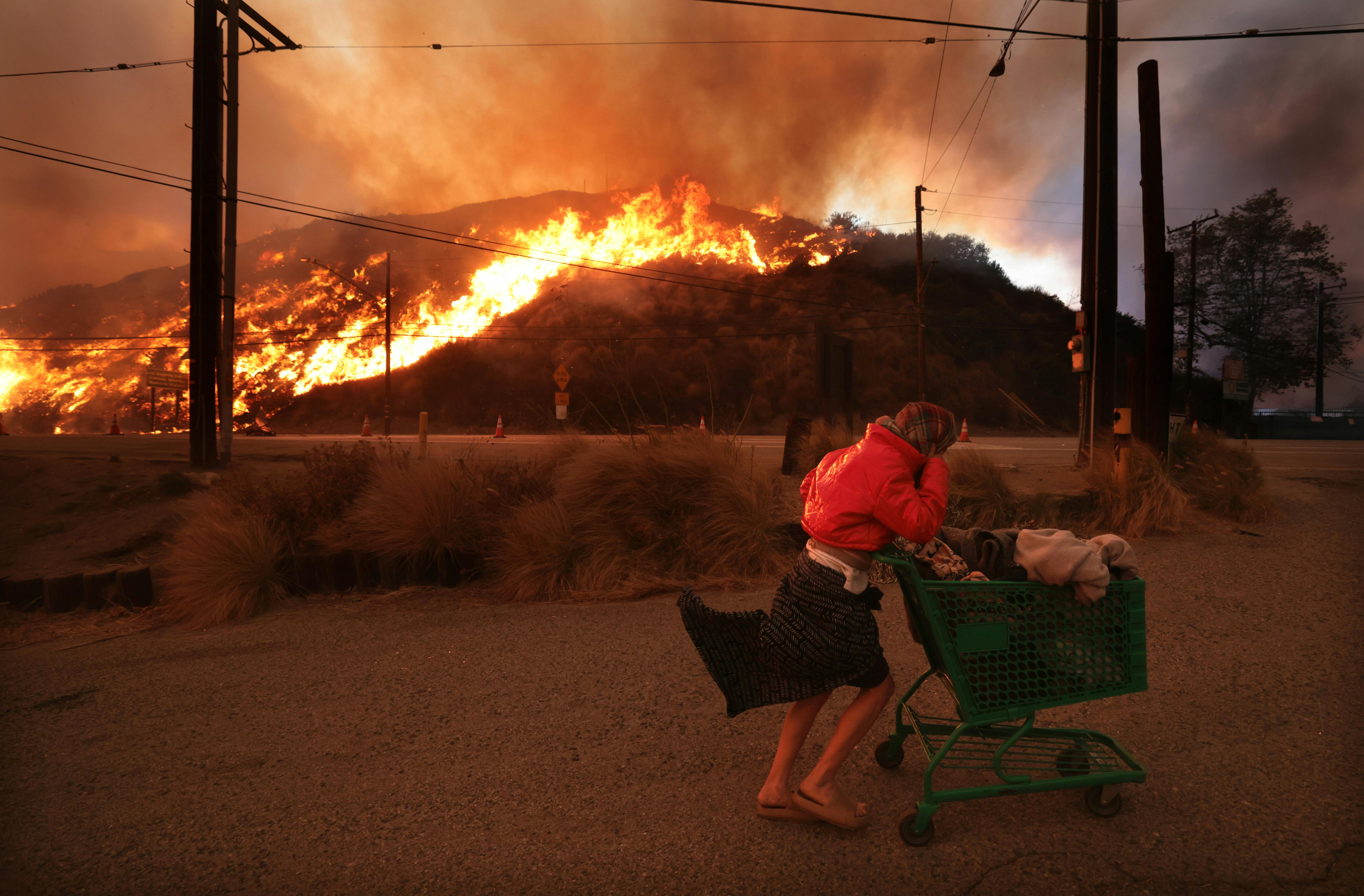A person pushes a cart of belongings with a burning hill in the background.