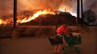 A person pushes a cart of belongings with a burning hill in the background.