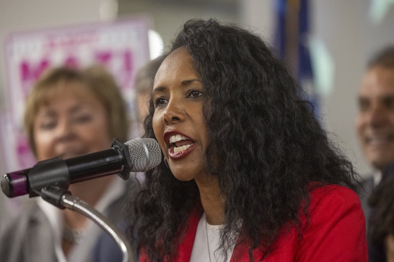 Mazi Pilip speaks at a mic. She is wearing a red blazer and has her curly hair down.