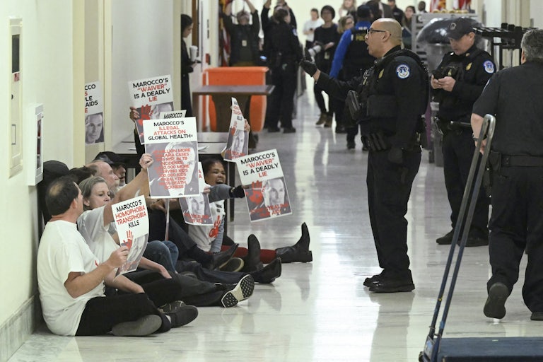 Protesters sit on the floor outside a door with signs like "Marocco's illegal attacks on global health." Capitol Police stand near them, with one officer yelling at them.