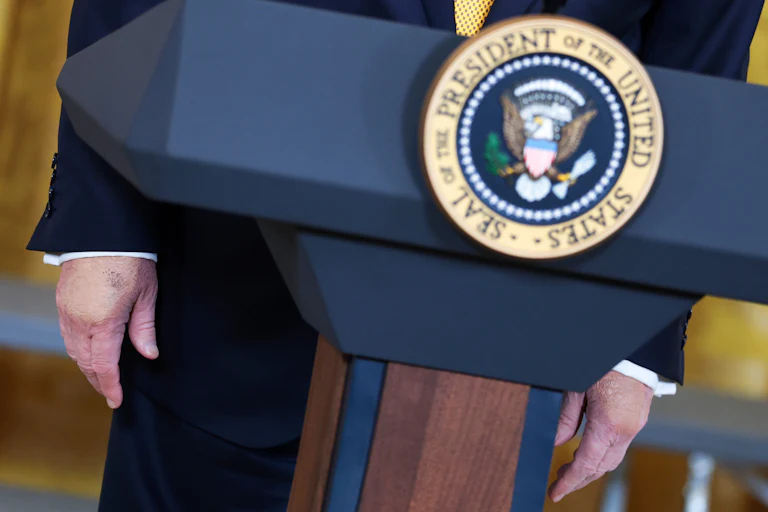 Donald Trump wears makeup on both hands. He stands at a podium