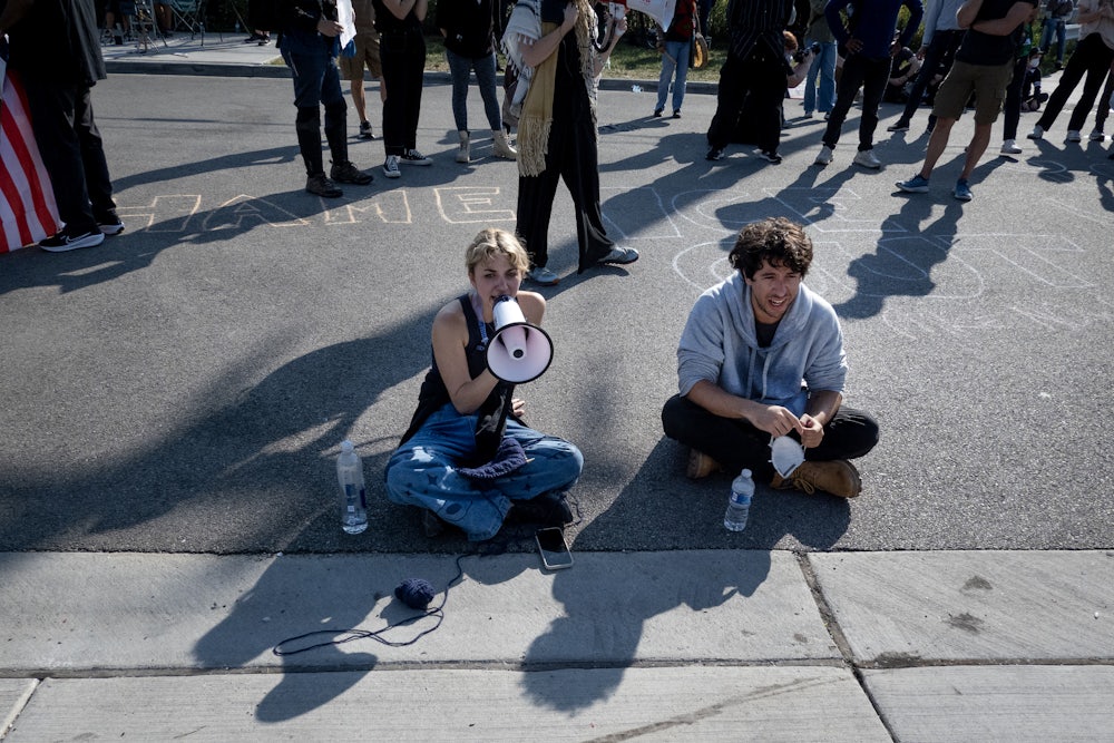 Kat Abughazaleh sits on the pavement and speaks into a megaphone. She is sitting next to another protester and in front of several others