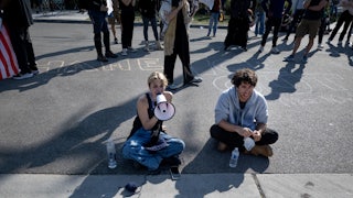 Kat Abughazaleh sits on the pavement and speaks into a megaphone. She is sitting next to another protester and in front of several others