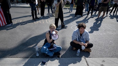 Kat Abughazaleh sits on the pavement and speaks into a megaphone. She is sitting next to another protester and in front of several others