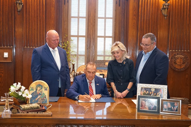 Missouri Governor Mike Kehoe signs HB 567, repealing the state's paid sick leave. In his office is a portrait of the Virgin Mary, a framed photo of him and his wife, and a large Blue Lives Matter flag. One white woman and two white men look down and smile as he signs the legislation.