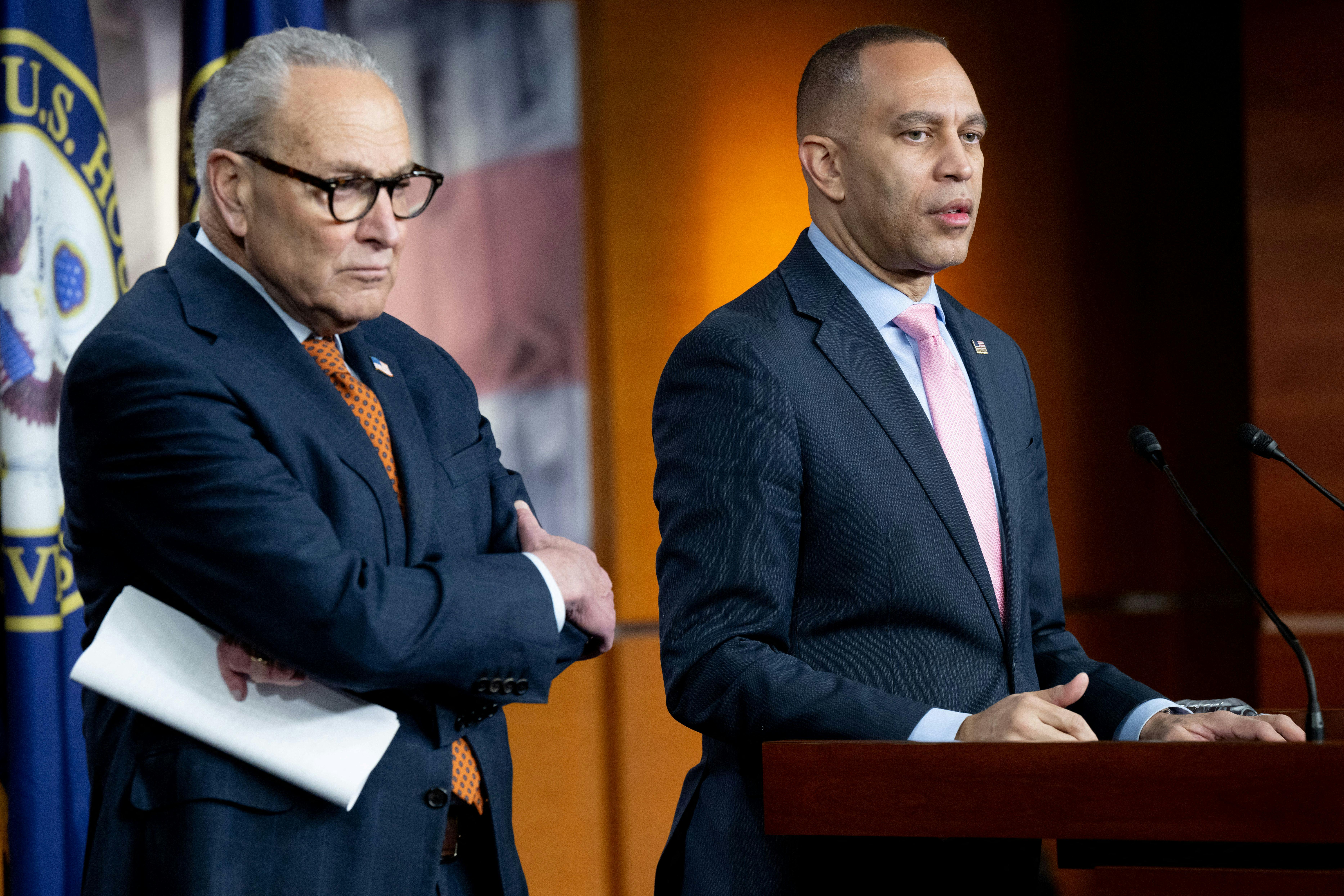 Senate Minority Leader Chuck Schumer crosses his arms, as House Minority Leader Hakeem Jeffries, beside him, speaks at a podium in the Capitol.