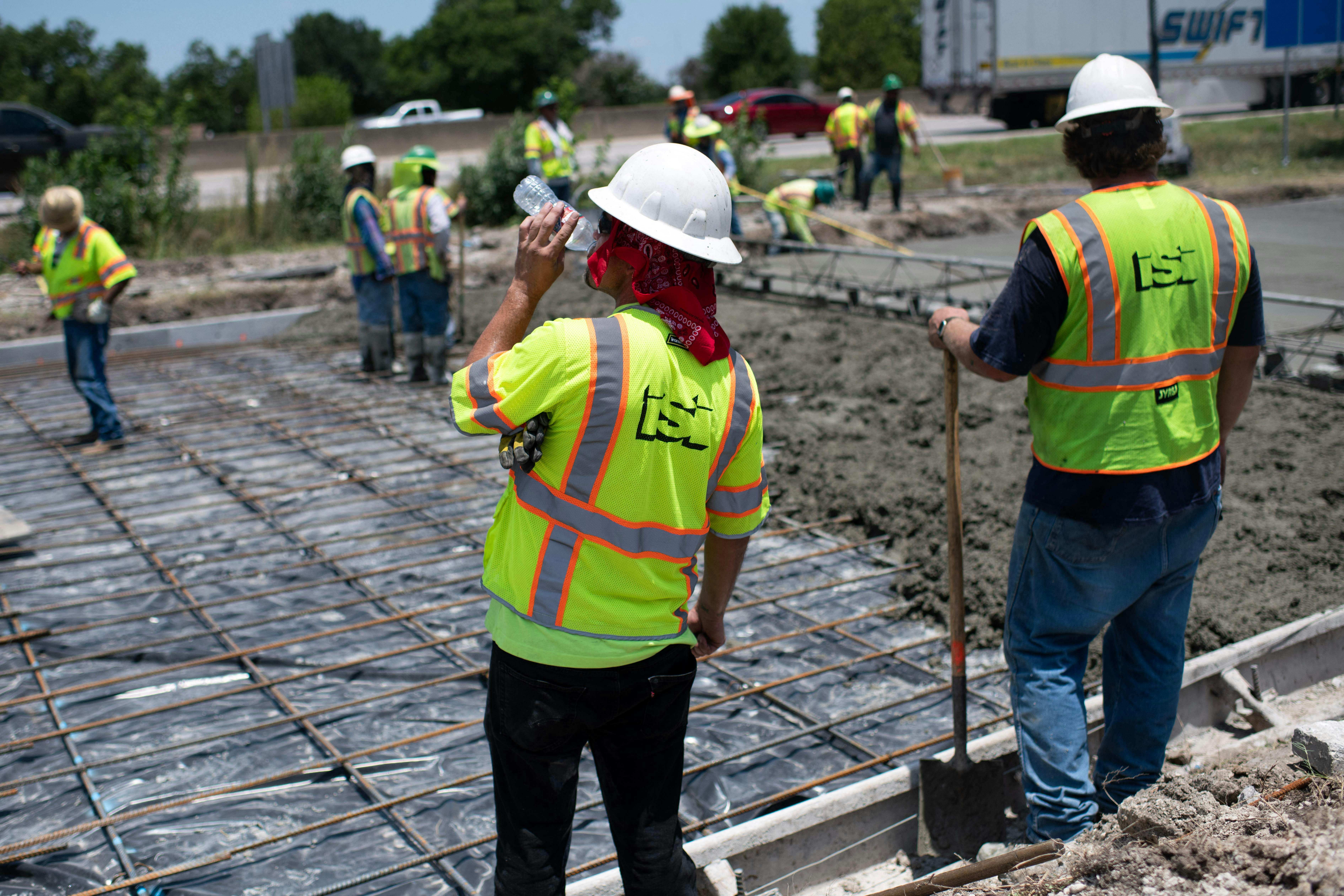 A man in a neon vest sips from a plastic water bottle; in the background, other people spread concrete on a road.