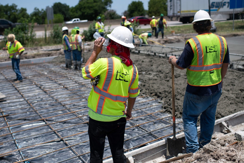 A man in a neon vest sips from a plastic water bottle; in the background, other people spread concrete on a road.