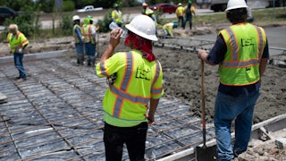 A man in a neon vest sips from a plastic water bottle; in the background, other people spread concrete on a road.