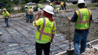 A man in a neon vest sips from a plastic water bottle; in the background, other people spread concrete on a road.