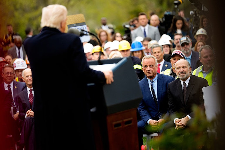 RFK Jr. sits in the audience, watching Trump, who stands at a podium facing away from the camera.