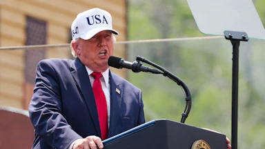 Donald Trump yelling at a podium while wearing a white USA cap