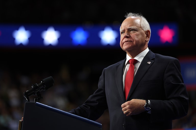 Tim Walz stares into the distance with a hand on his suit button while he stands at a lectern