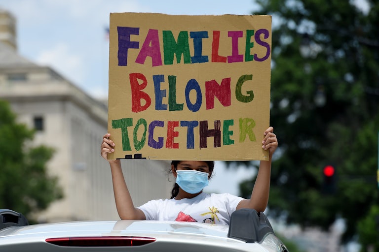 A child sticking out of a car sunroof and wearing a face mask holds a sign that reads "Families Belong Together."