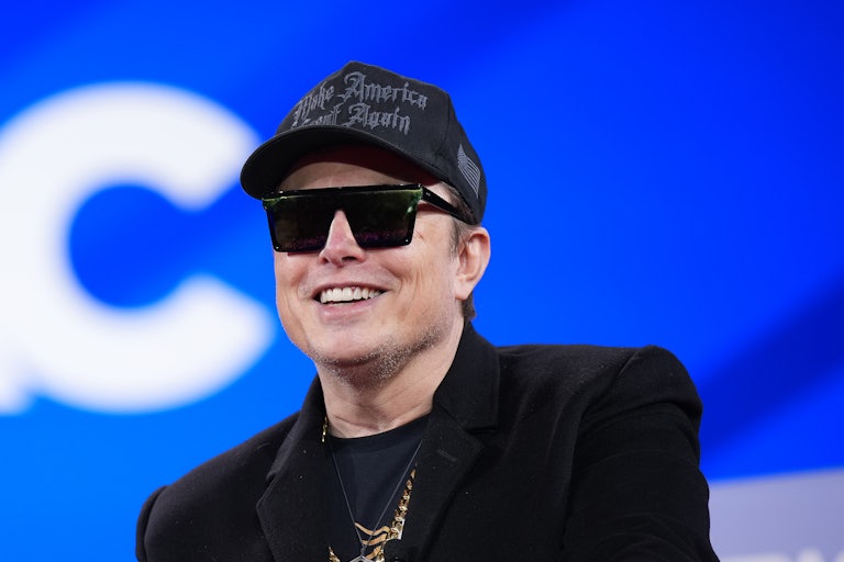 Elon Musk smiles while wearing sunglasses, all black, and a black Make America Great Again cap. (He is indoors.)