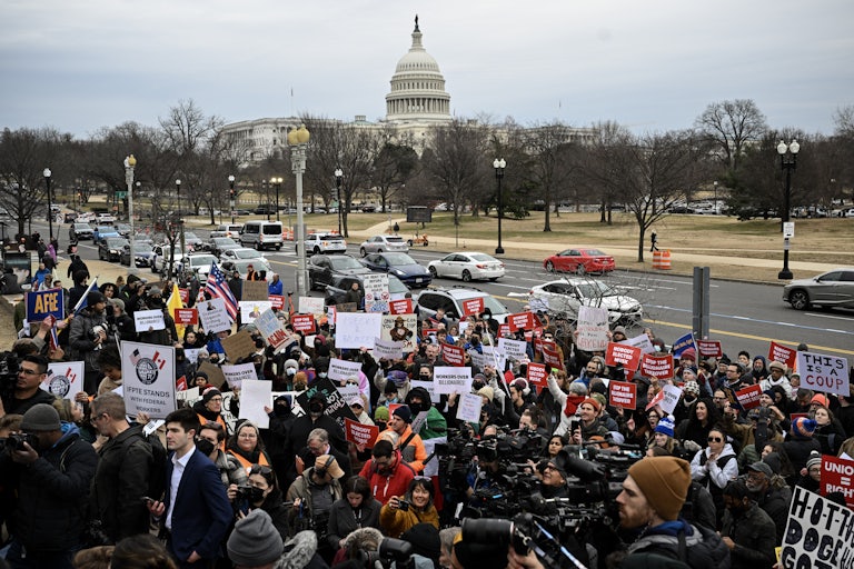 Protestors hold signs against Donald Trump and Elon Musk across the street from the U.S. Capitol building, which is surrounded by trees.