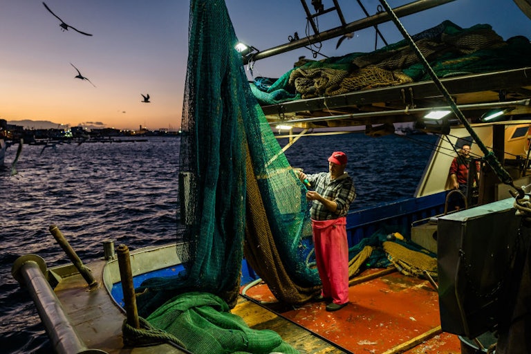A person stands on a boat in the middle of the water holding onto a net that is hoisted above him.