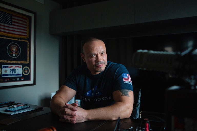 Sitting in a dark room Dan Bongino leans forward while sitting behind a table and clasping his hands