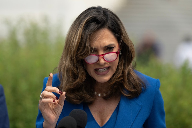 Representative Maria Salazar wears a blue blazer and red glasses. She is speaking outside and gesturing with her index finger raised.