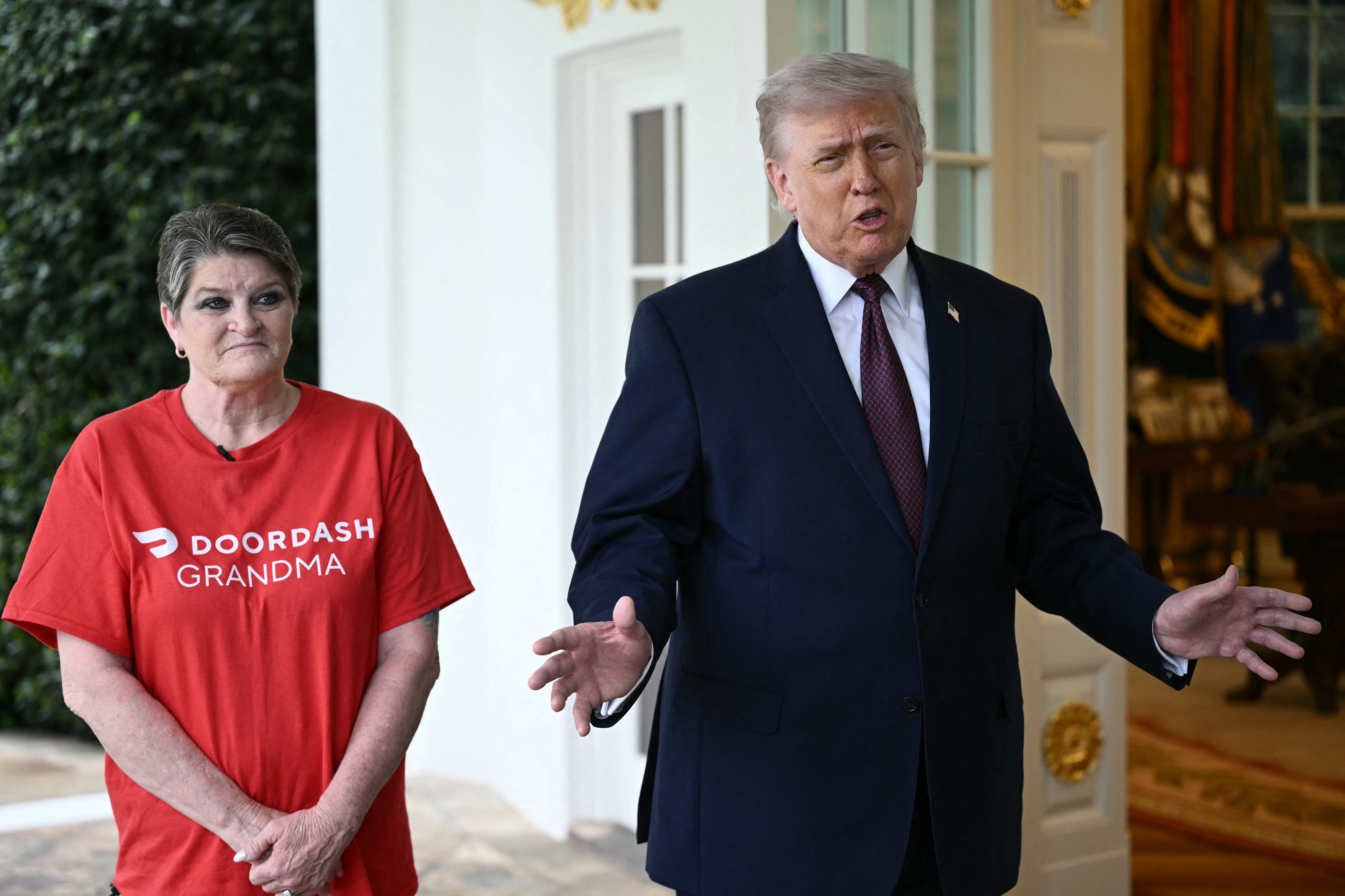Sharon Simmons wearing a "DoorDash Grandma" t-shirt stands next to President Trump as he speaks outside the Oval Office of the White House.