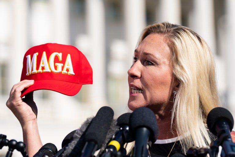 Marjorie Taylor Greene holds a red MAGA cap and speaks before several mics outside the Capitol.